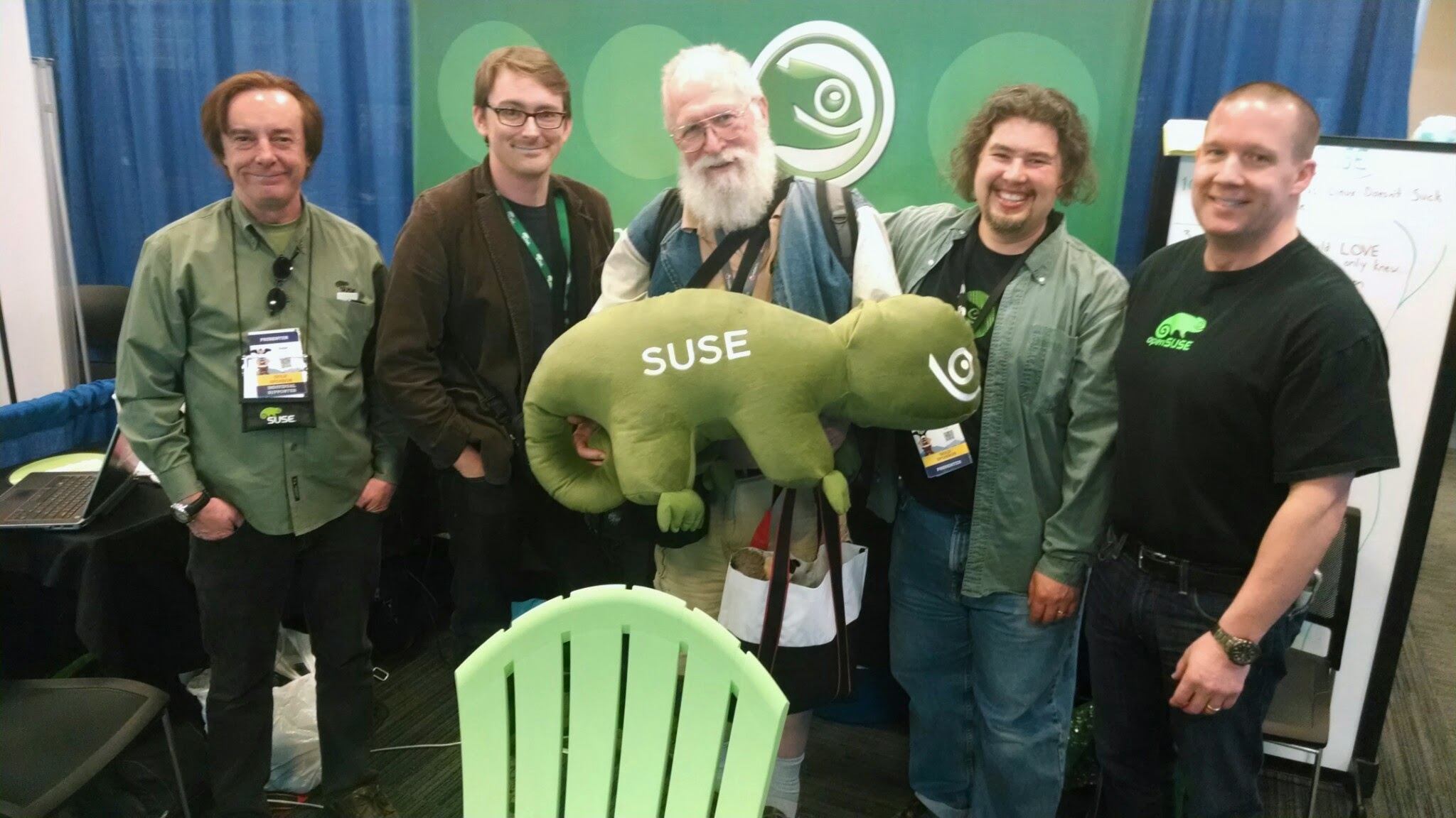 A group of community members at a SUSE booth at LinuxFest Northwest 2014, including Jon "maddog" Hall holding a plush SUSE chameleon mascot